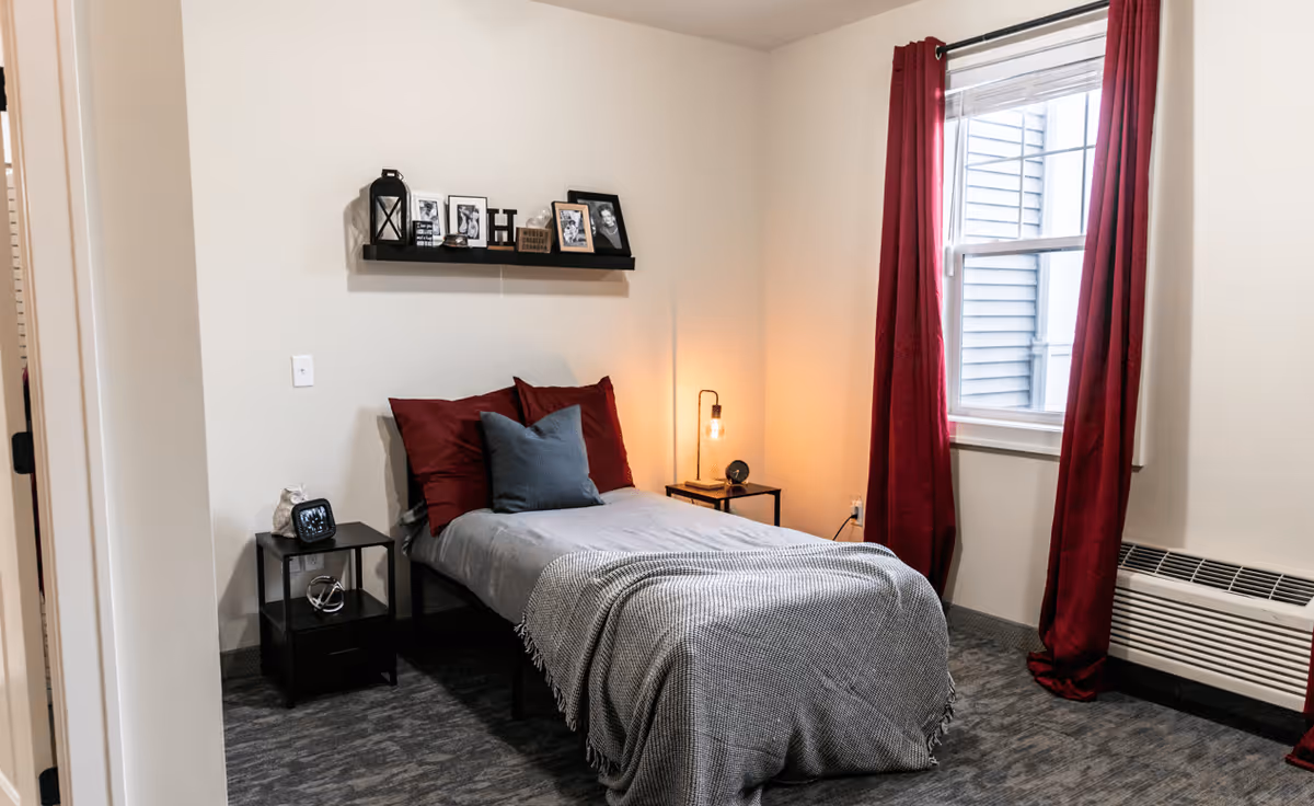A cozy bedroom with a single bed made with gray and burgundy bedding, a small black nightstand with a clock and decorative items, a lit bedside lamp, a window with burgundy curtains, and a wall shelf displaying framed photos and decorative pieces.