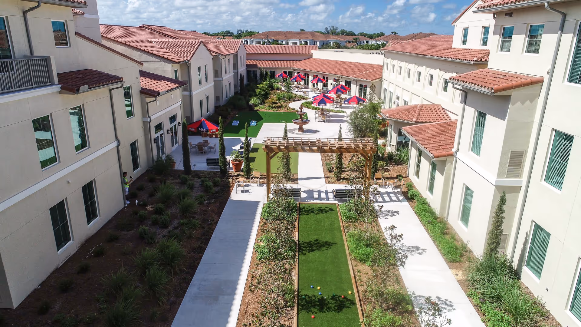 Outdoor courtyard area at Tuscan Gardens of Delray Beach featuring a bocce ball court with colored balls, a wooden pergola, landscaped garden beds, paved walkways, a central fountain, and several tables with red and blue umbrellas. The courtyard is surrounded by beige buildings with red tile roofs under a partly cloudy sky.