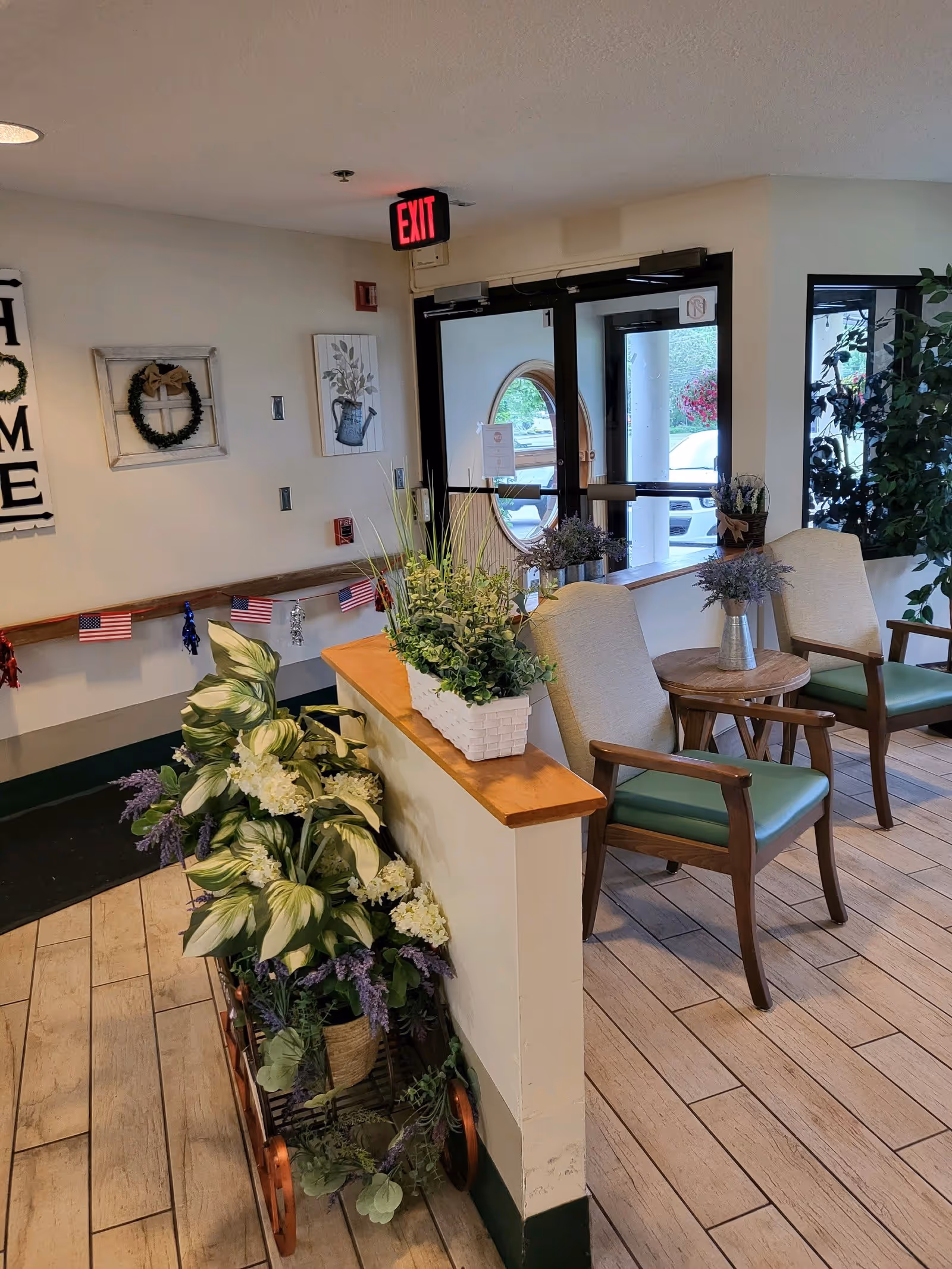 Interior view of a senior living facility entrance area with two wooden chairs with green cushions, a small round wooden table with a vase of flowers, a decorative cart filled with green and white plants, and a wooden ledge with a white planter of greenery. The entrance door has a round window, and there is an illuminated red exit sign above it. The walls are decorated with a 'HOME' sign, a wreath, and a framed picture of a watering can. Small American flags and red, white, and blue decorations hang along the wall.