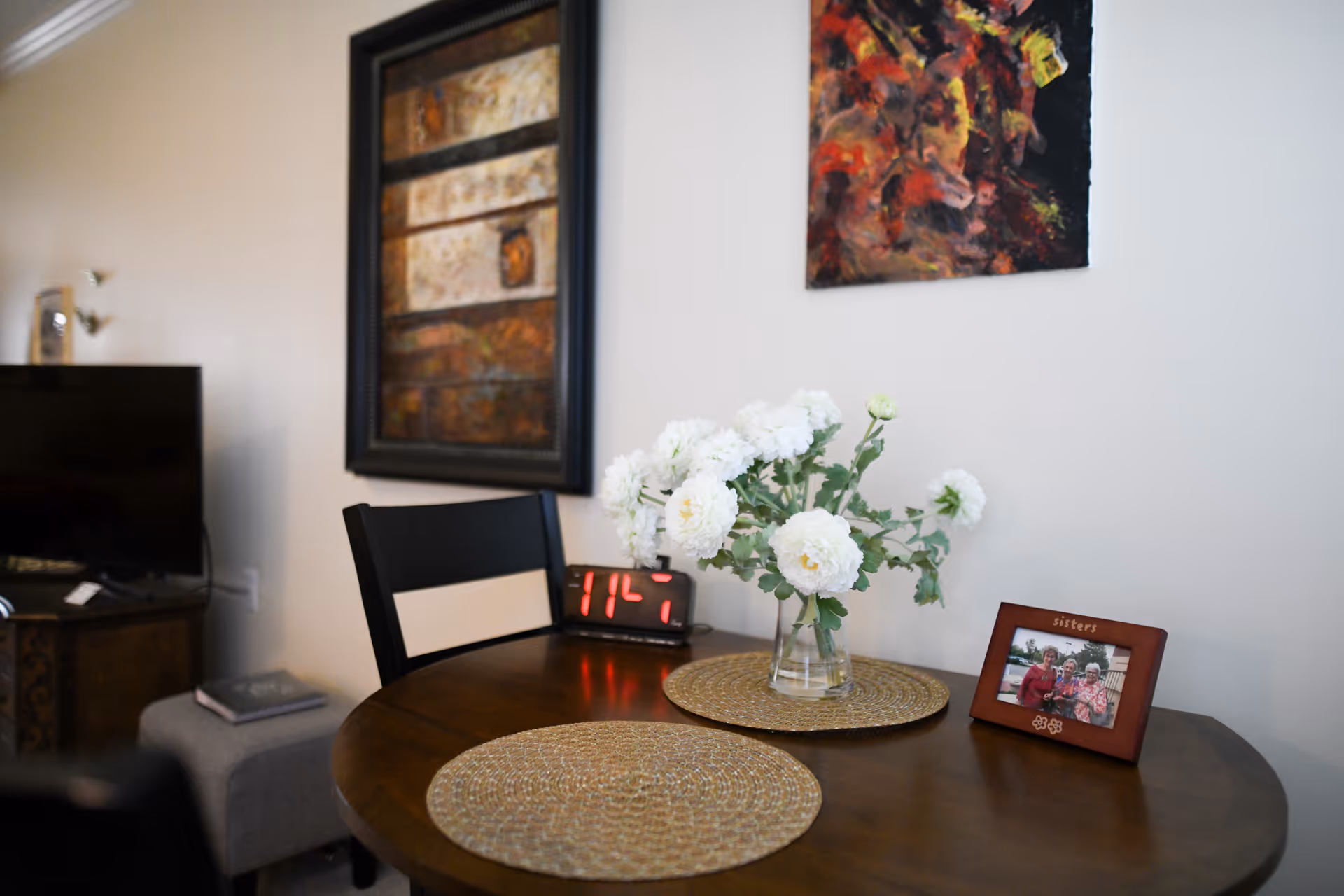 A small round wooden table with two woven placemats, a glass vase with white flowers, and a framed photo labeled 'sisters'. Behind the table is a black chair, a digital clock showing 11:47, and a wall with two pieces of artwork. A television and a small ottoman with a book are visible in the background.