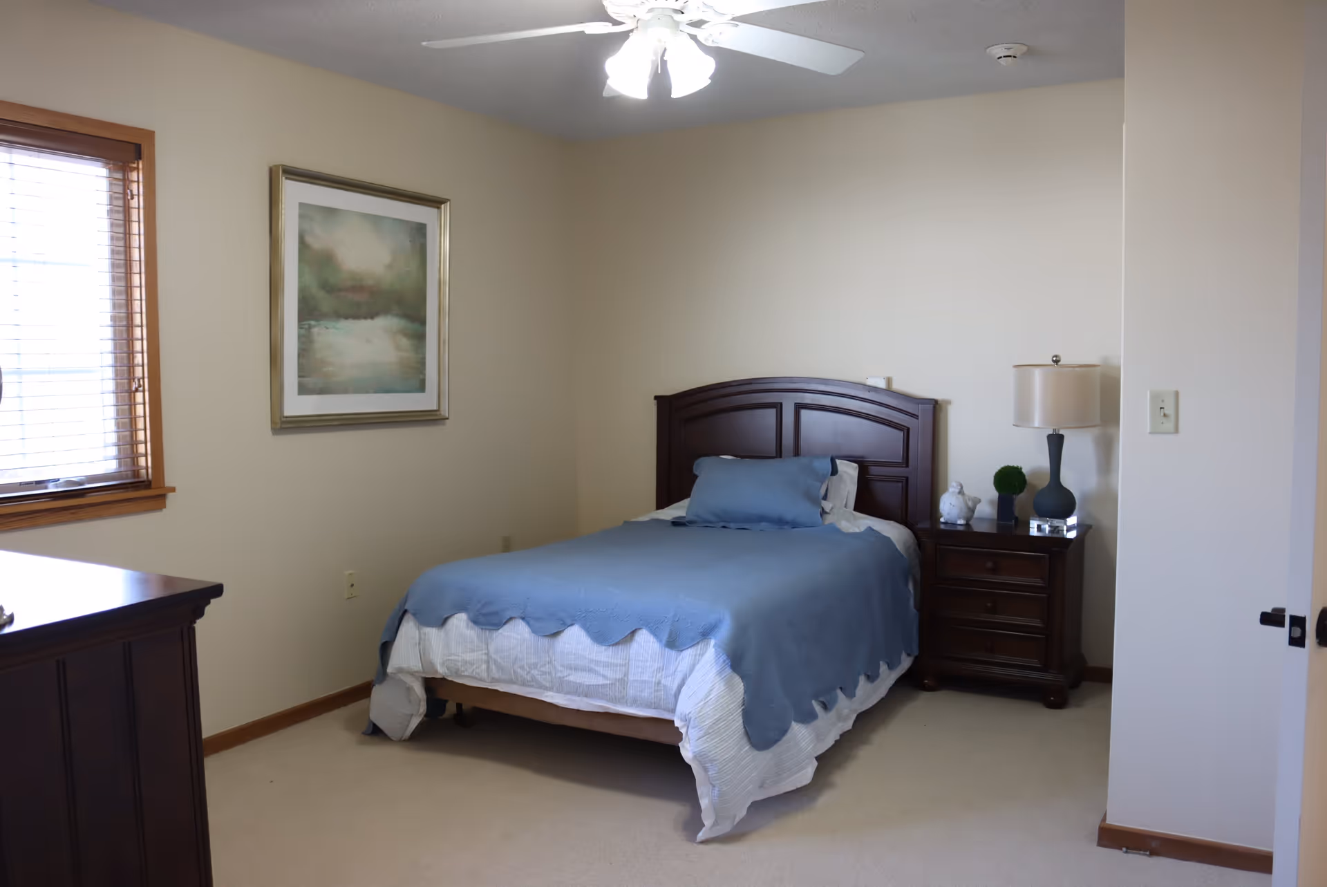 A simple bedroom with a single bed covered with a blue blanket and a matching pillow. The bed has a dark wooden headboard. Next to the bed is a dark wooden nightstand with a lamp, a small plant, and a decorative bird. There is a framed painting on the beige wall and a window with wooden blinds letting in natural light. A ceiling fan with lights is on the ceiling.