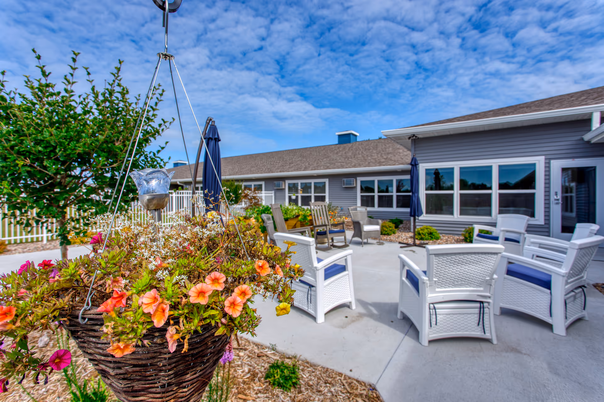 Outdoor patio area at The Cottages at Lake Park Senior Living & Memory Care featuring white wicker chairs with blue cushions arranged in a circle on a concrete surface, two closed navy blue umbrellas, a hanging basket with colorful flowers in the foreground, and a gray building with multiple windows in the background under a partly cloudy sky.