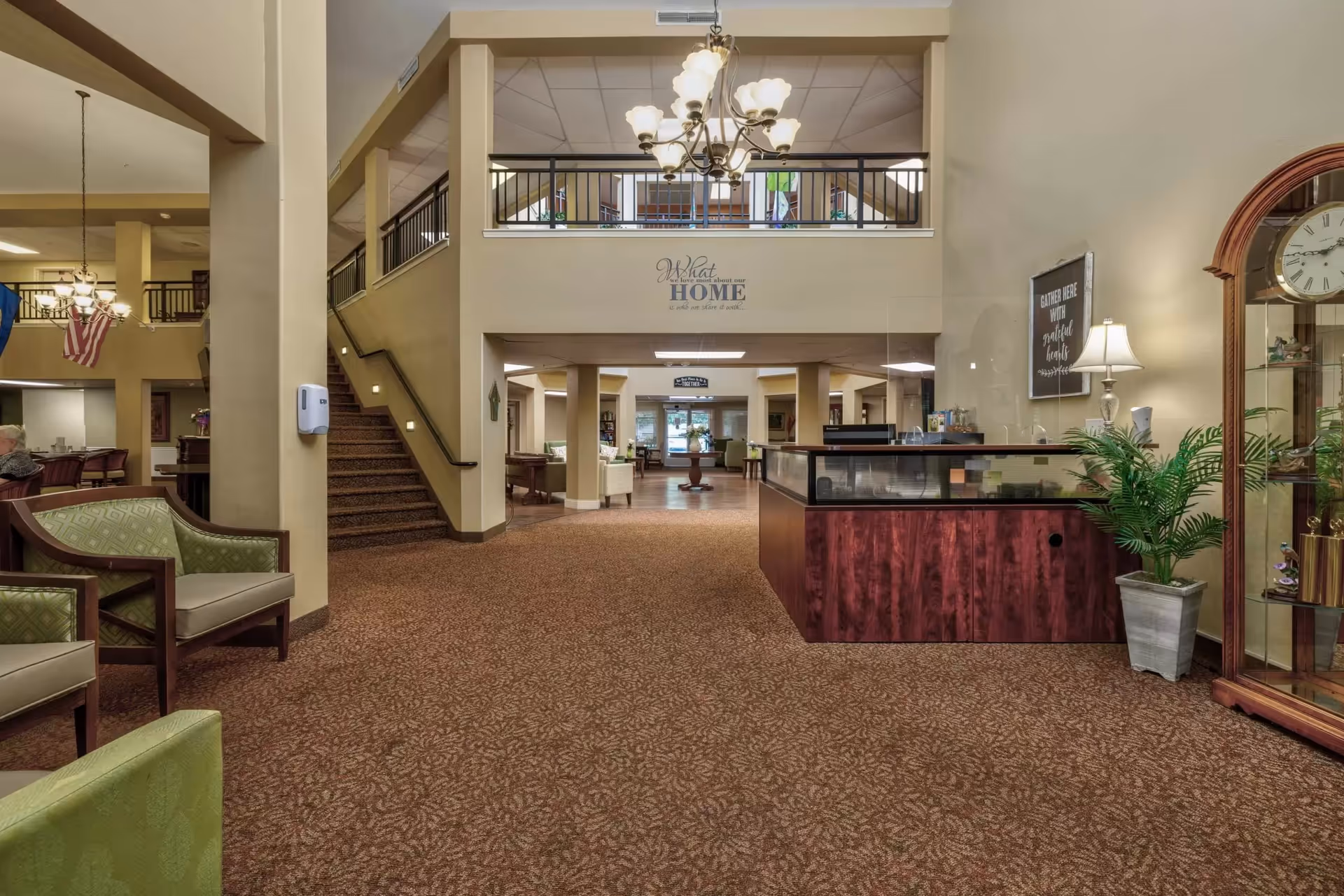 Spacious residential lobby with carpeted floor, seating areas, a wooden reception desk, a central chandelier, and a staircase leading to an upper balcony.