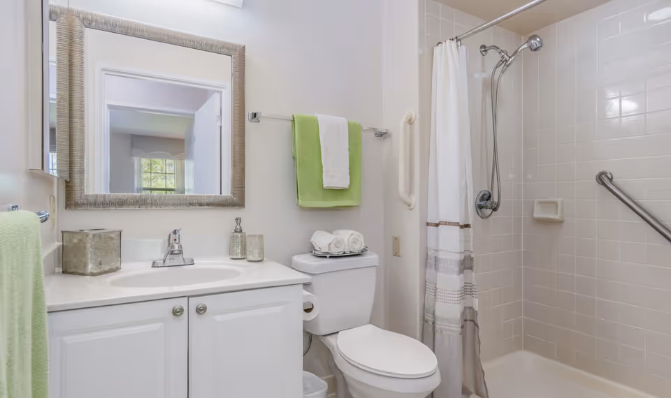 Bathroom featuring a white vanity with mirror, toilet topped with rolled towels, green towels, and a tiled walk-in shower with grab bars.