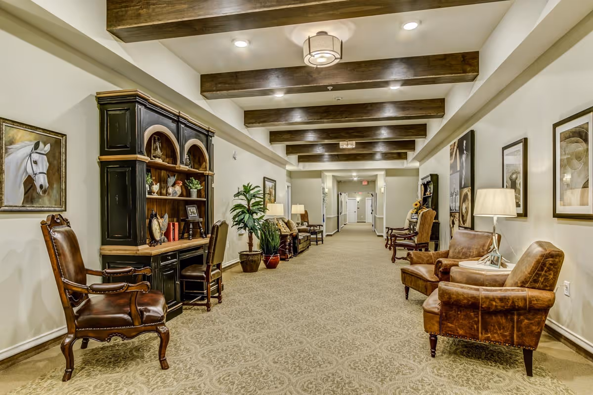 A long, well-lit hallway in an assisted living facility with beige patterned carpet, wooden ceiling beams, and cream-colored walls. The hallway is furnished with various chairs, a black wooden cabinet with shelves holding decorative items, potted plants, framed artwork on the walls, and table lamps providing warm lighting.