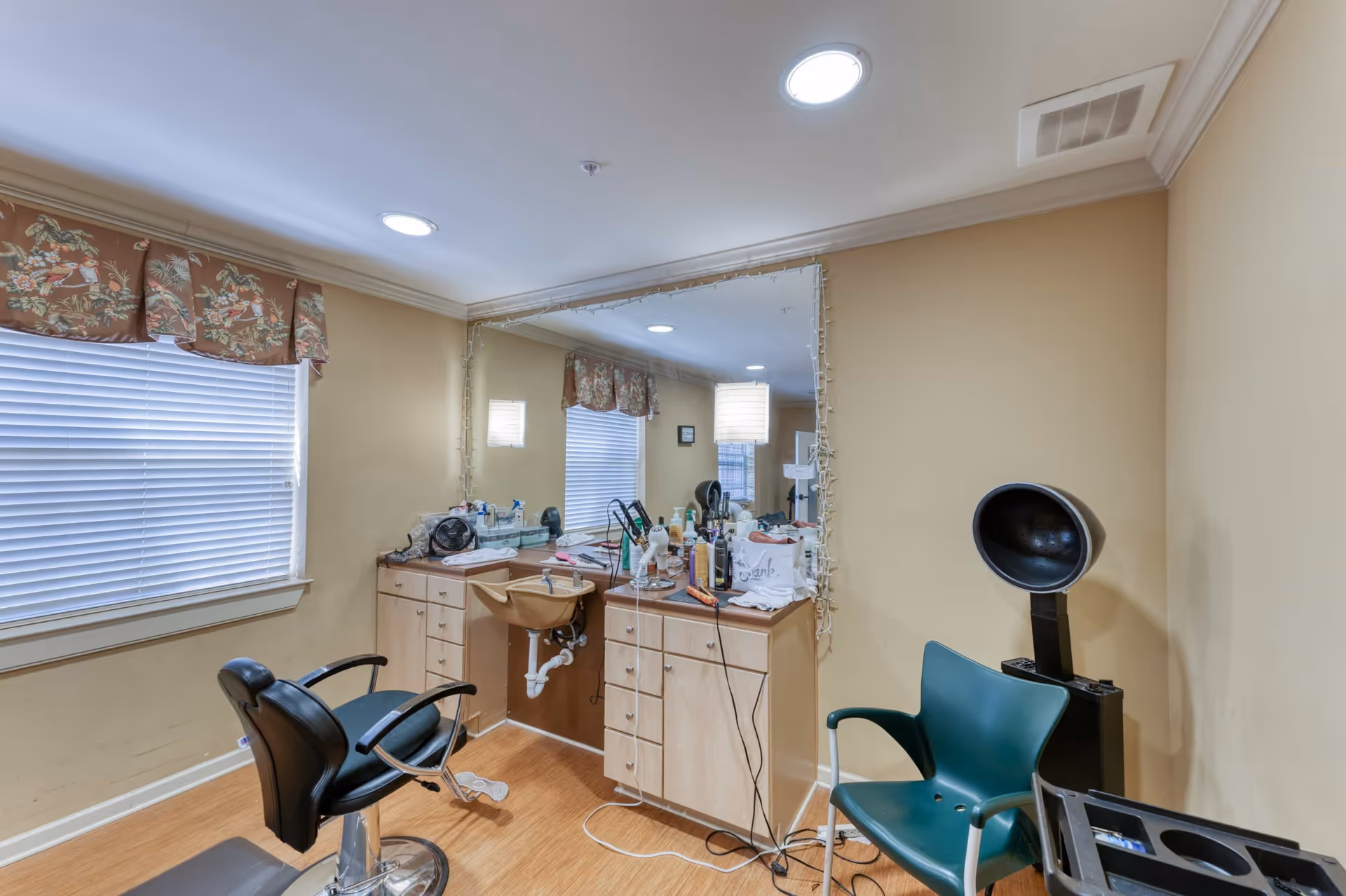Interior view of a small hair salon or beauty station with a black salon chair, a green plastic chair, a hair dryer hood, a large mirror with string lights around it, and a countertop cluttered with hair care products and tools. The room has beige walls, a window with blinds and a floral valance, and recessed ceiling lights.
