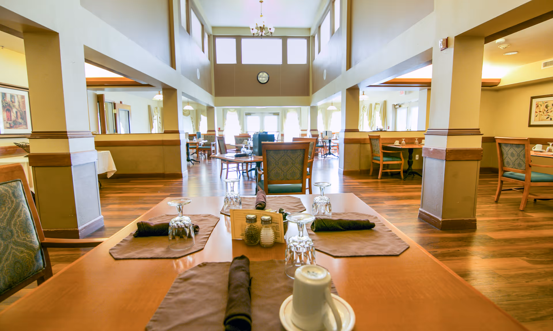 Bright, spacious dining room with wooden tables set with napkins, glassware and chairs beneath a high ceiling and large windows.