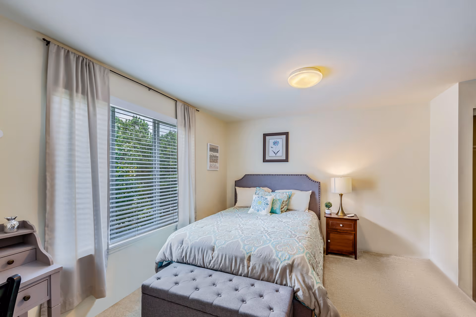 A cozy bedroom with a large window covered by white curtains and blinds. The bed has a gray upholstered headboard and is made with a patterned comforter and several decorative pillows. A wooden nightstand with a lamp and a small plant is beside the bed. There is a gray tufted bench at the foot of the bed and a small desk with drawers on the left side of the room. The walls are painted light beige and decorated with framed artwork.