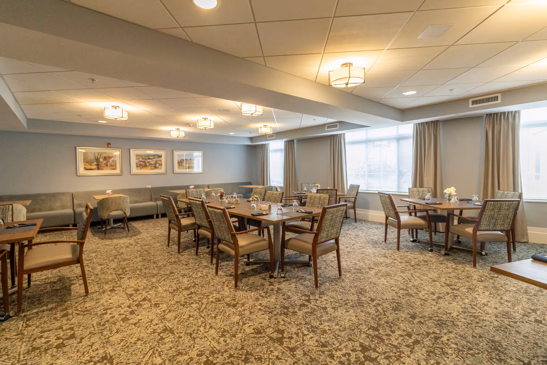 A spacious dining room with multiple wooden tables and cushioned chairs arranged neatly. The room has large windows with beige curtains allowing natural light to fill the space. The walls are decorated with framed artwork, and the ceiling has modern light fixtures. The floor is covered with a patterned carpet.