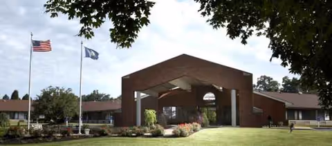 Brick senior care facility entrance with flagpoles and a manicured lawn.