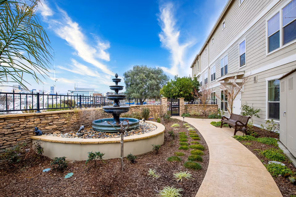 Outdoor garden area at Oakmont of Mariner Point featuring a multi-tiered black fountain surrounded by rocks and plants, a curved concrete pathway, a wooden bench, and a beige building with multiple windows under a blue sky with wispy clouds.