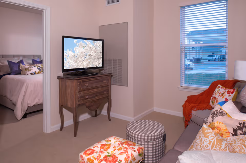 A cozy living room with a TV on a wooden dresser, patterned ottomans and a sofa with colorful pillows, and a view into a bedroom.