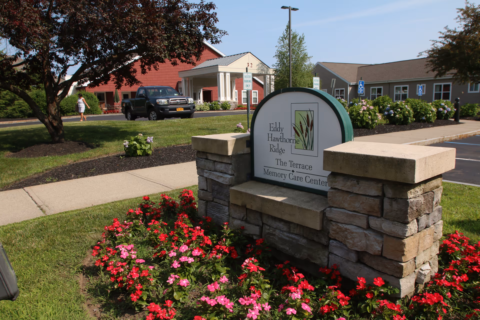Stone entrance sign for Eddy Hawthorne Ridge memory care surrounded by flowers with the facility buildings and a parked truck behind.