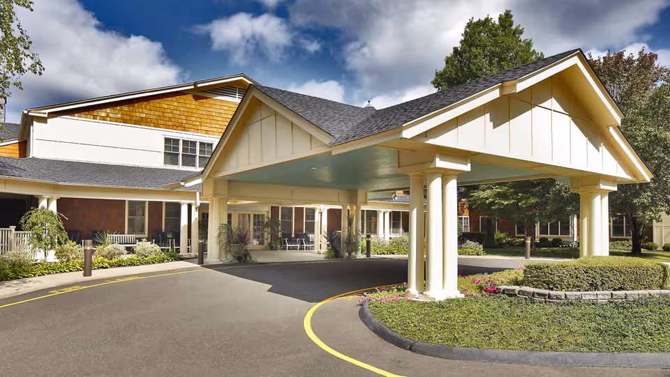 Entrance area of a senior living facility with a covered drop-off zone supported by white pillars, surrounded by landscaped greenery and trees under a partly cloudy sky.