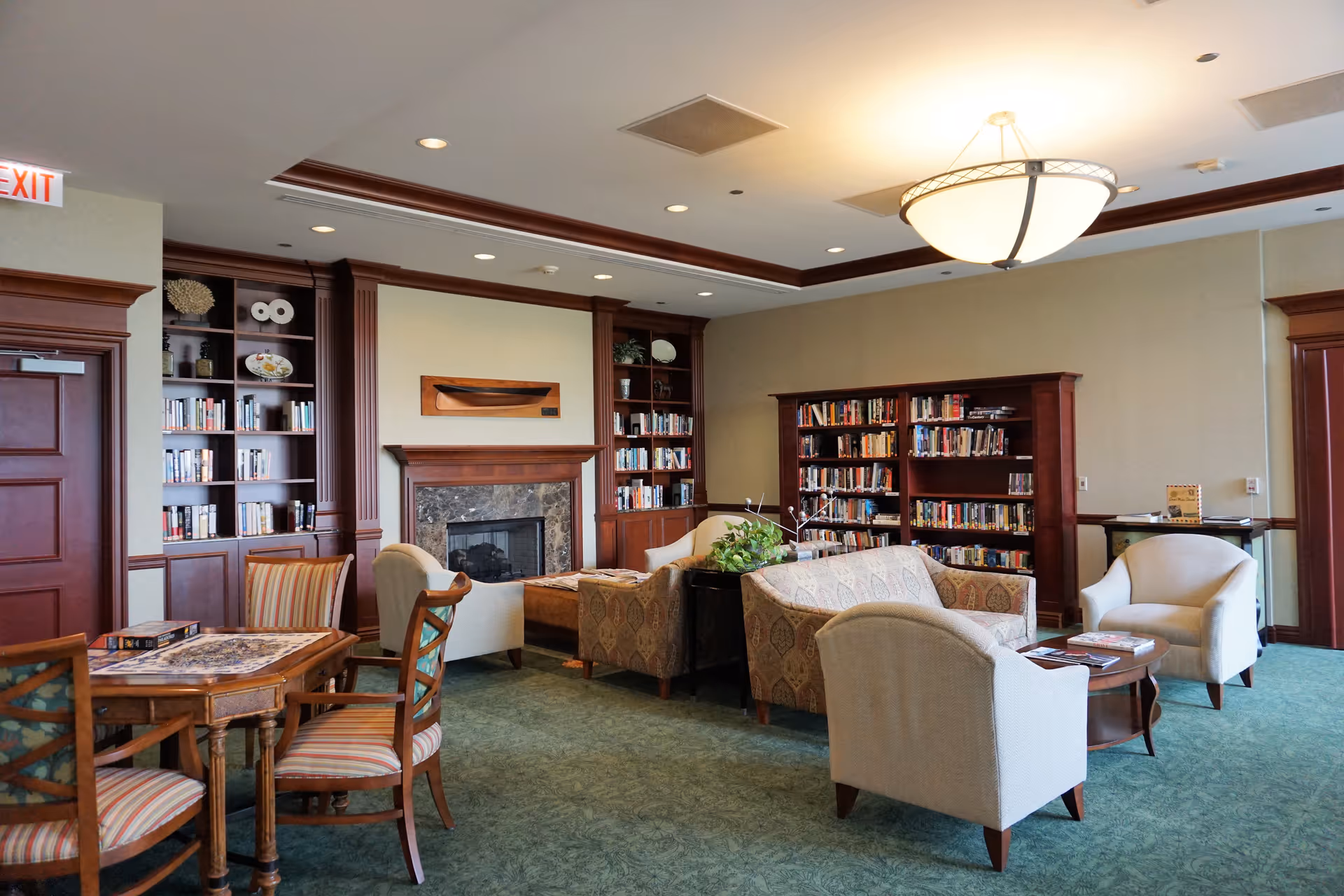 A cozy library or lounge area with bookshelves filled with books, comfortable armchairs and sofas arranged around a coffee table, a fireplace with a decorative wooden mantle, and a small table with chairs in the foreground. The room has green carpet, beige walls, and a ceiling light fixture.