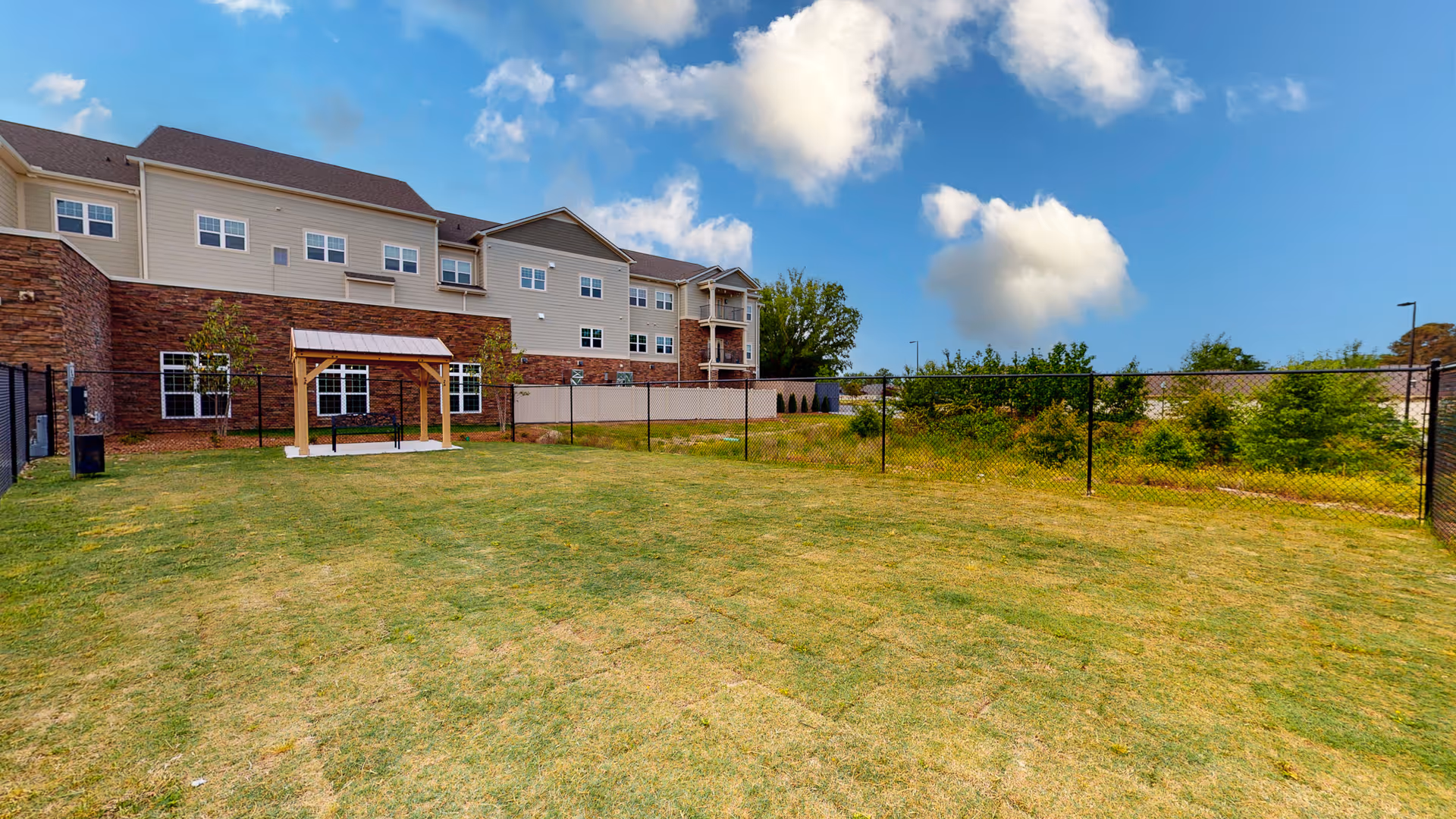 Outdoor fenced grassy area with a small wooden pergola and bench, adjacent to a multi-story building with beige siding and stone accents under a blue sky with scattered clouds.
