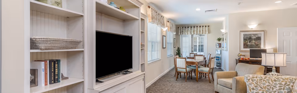 Interior view of a senior living facility common area with a built-in white entertainment center holding a flat-screen TV and shelves with books and decorative items. The room has large windows with floral valances, a wooden dining table with four chairs, an armchair, a side table with a lamp, and a desk with a computer and framed artwork on the wall.