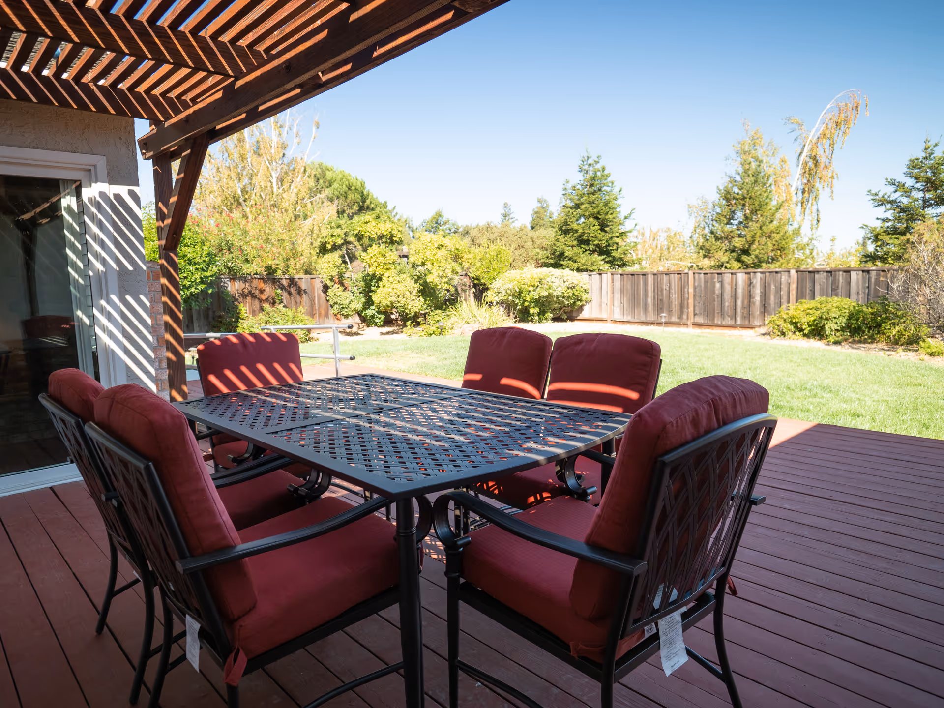 A shaded wooden deck patio with a metal dining table and six red-cushioned chairs under a pergola overlooking a grassy backyard.
