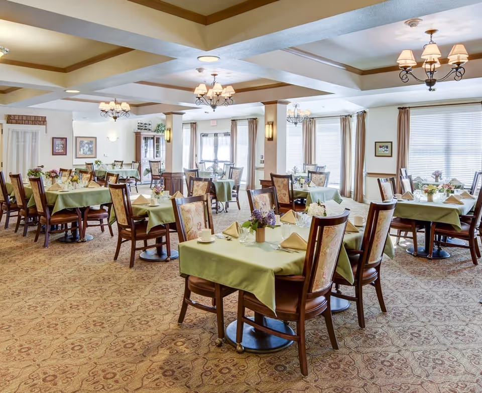 A spacious dining room with multiple tables covered in green tablecloths, each set with napkins, cups, and floral centerpieces. The room features patterned carpet, large windows with beige curtains, and decorative ceiling lights.