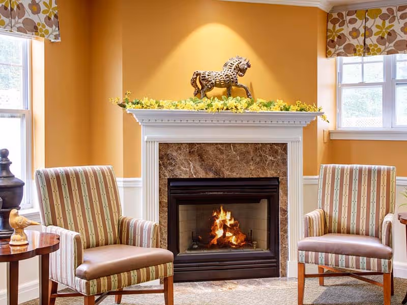 A cozy living room area with two striped armchairs facing a lit fireplace with a marble surround. Above the fireplace mantel is a decorative horse sculpture and a garland of yellow flowers. The walls are painted a warm yellow, and there are two windows with patterned valances on either side of the fireplace.