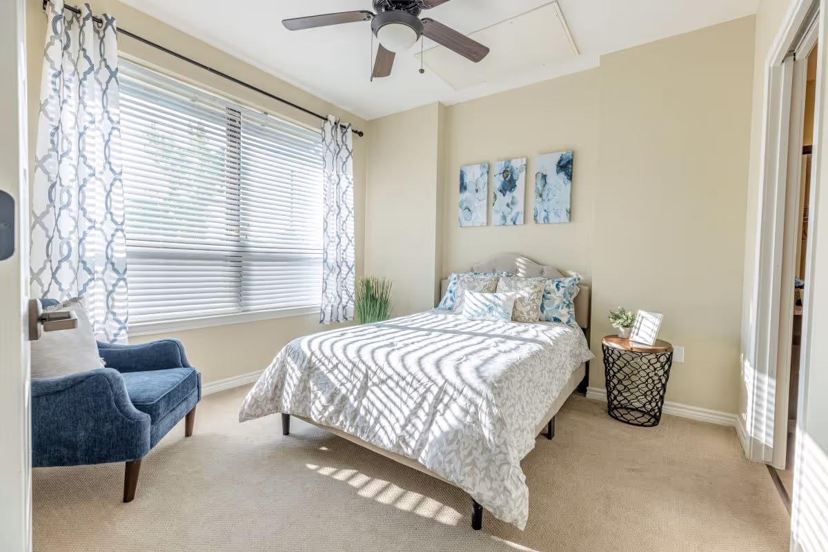 A bright bedroom with a bed covered in white and blue patterned bedding, a blue upholstered chair near a large window with white blinds and patterned curtains, a ceiling fan, and a small round side table with a plant and a framed photo.