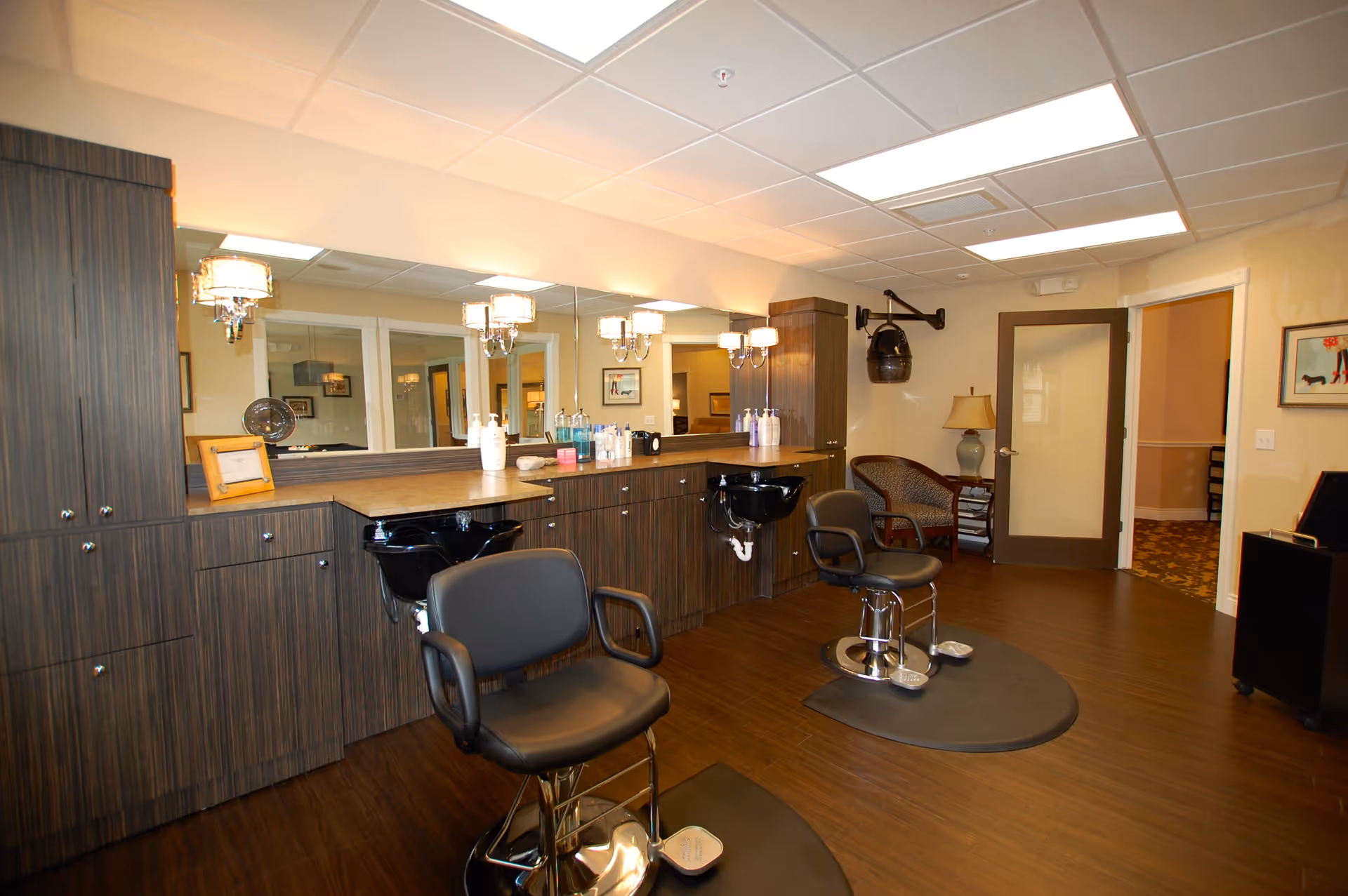 Interior view of a hair salon area in a senior living facility with two black salon chairs in front of a long counter with sinks and various hair care products. The room has wooden flooring, a large mirror with wall-mounted lights, and a seating area with a chair and lamp near a door.
