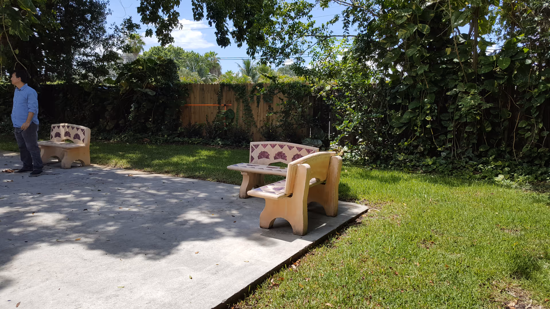 Outdoor patio area with concrete flooring and two sets of stone benches and tables. There is green grass surrounding the patio and a wooden fence with dense greenery and trees in the background. A person wearing a blue shirt and dark pants is standing on the left side near one of the benches.