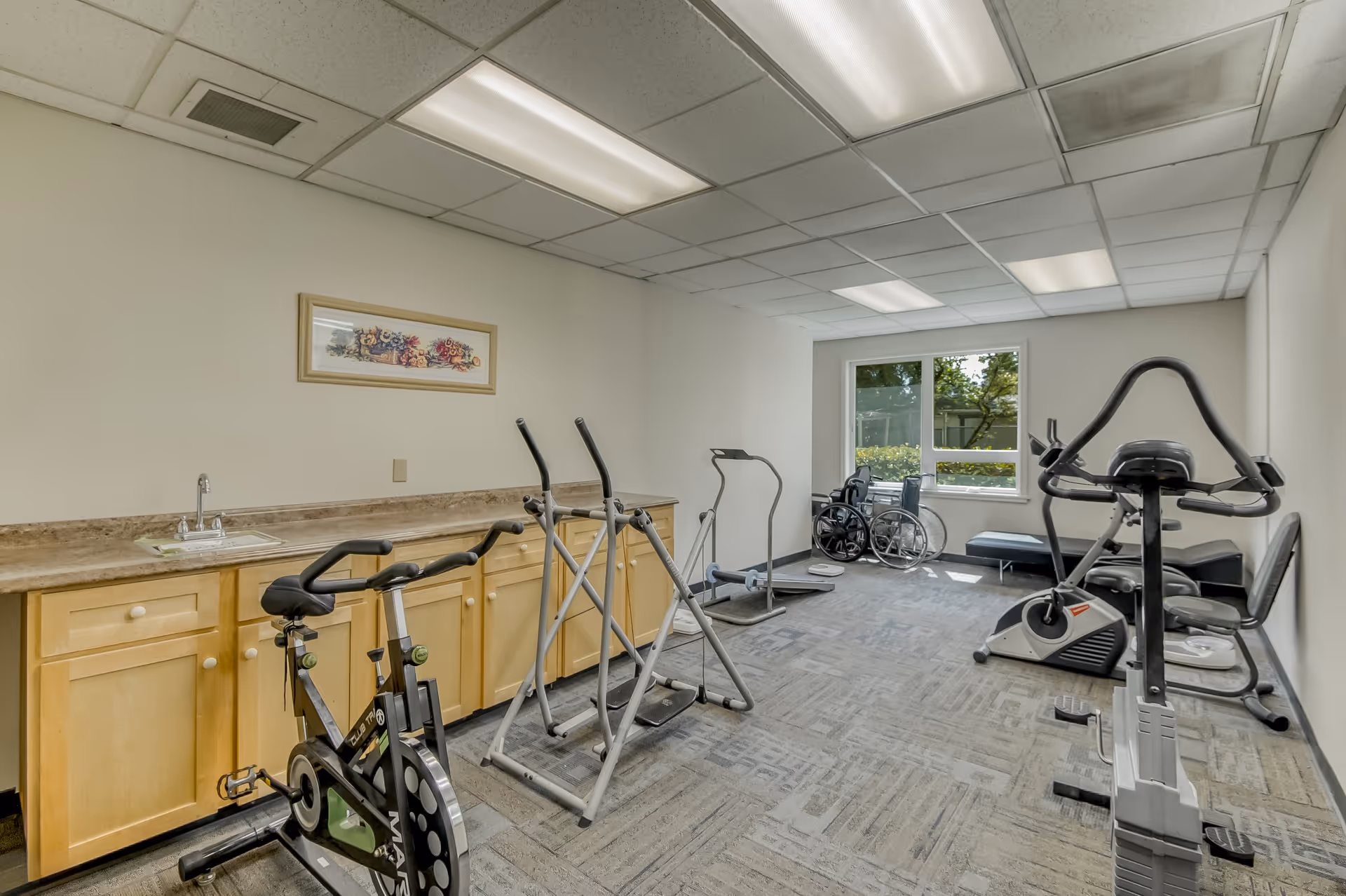 A small fitness room with exercise equipment including stationary bikes, elliptical machines, and a treadmill. There is a countertop with a sink and cabinets along one wall, a framed floral picture above the countertop, and a window letting in natural light. A wheelchair and a bench are placed near the window.