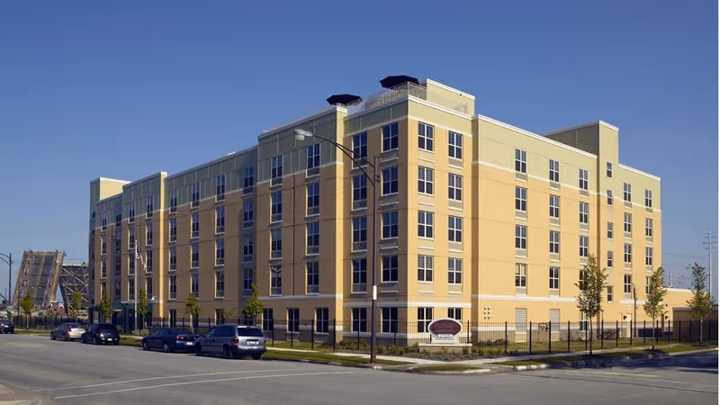Front exterior of a multi-story beige senior living building with parked cars and a fenced lawn under a clear blue sky.