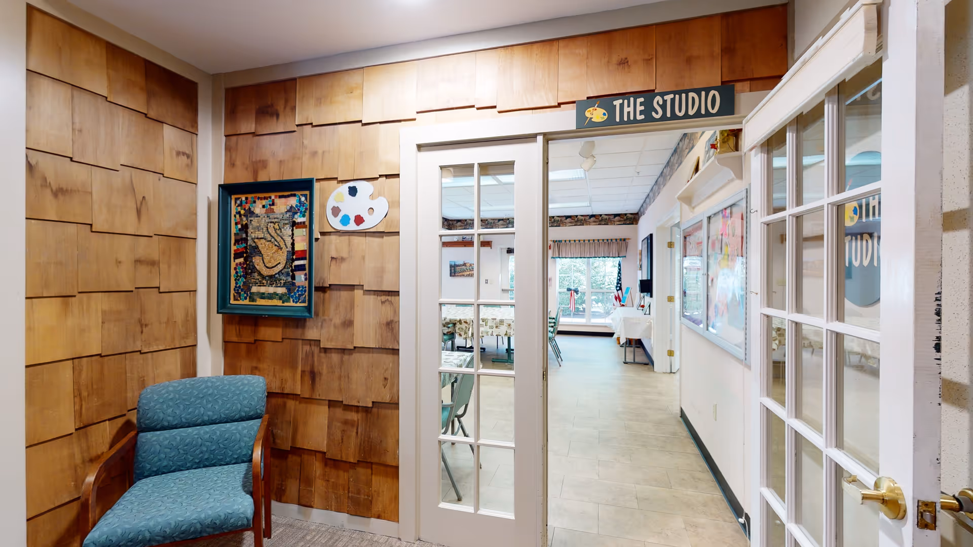 Interior view of a room labeled 'The Studio' with wooden panel walls, a blue upholstered chair, and glass-paned double doors leading to a larger room with tables and chairs, bulletin boards, and windows letting in natural light.