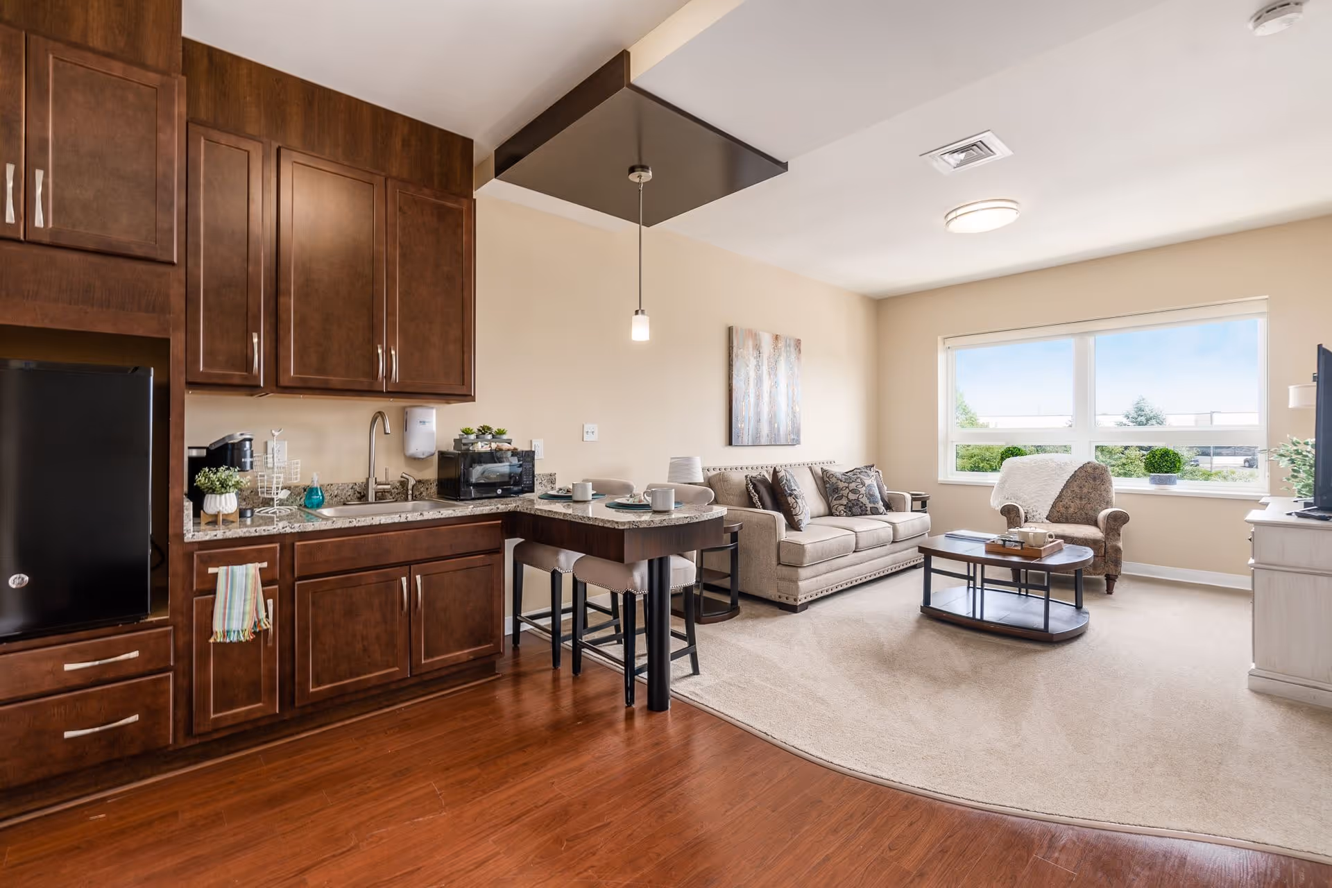 Open living room and kitchenette with dark wood cabinets, a sofa and armchair, coffee table, and a large window.