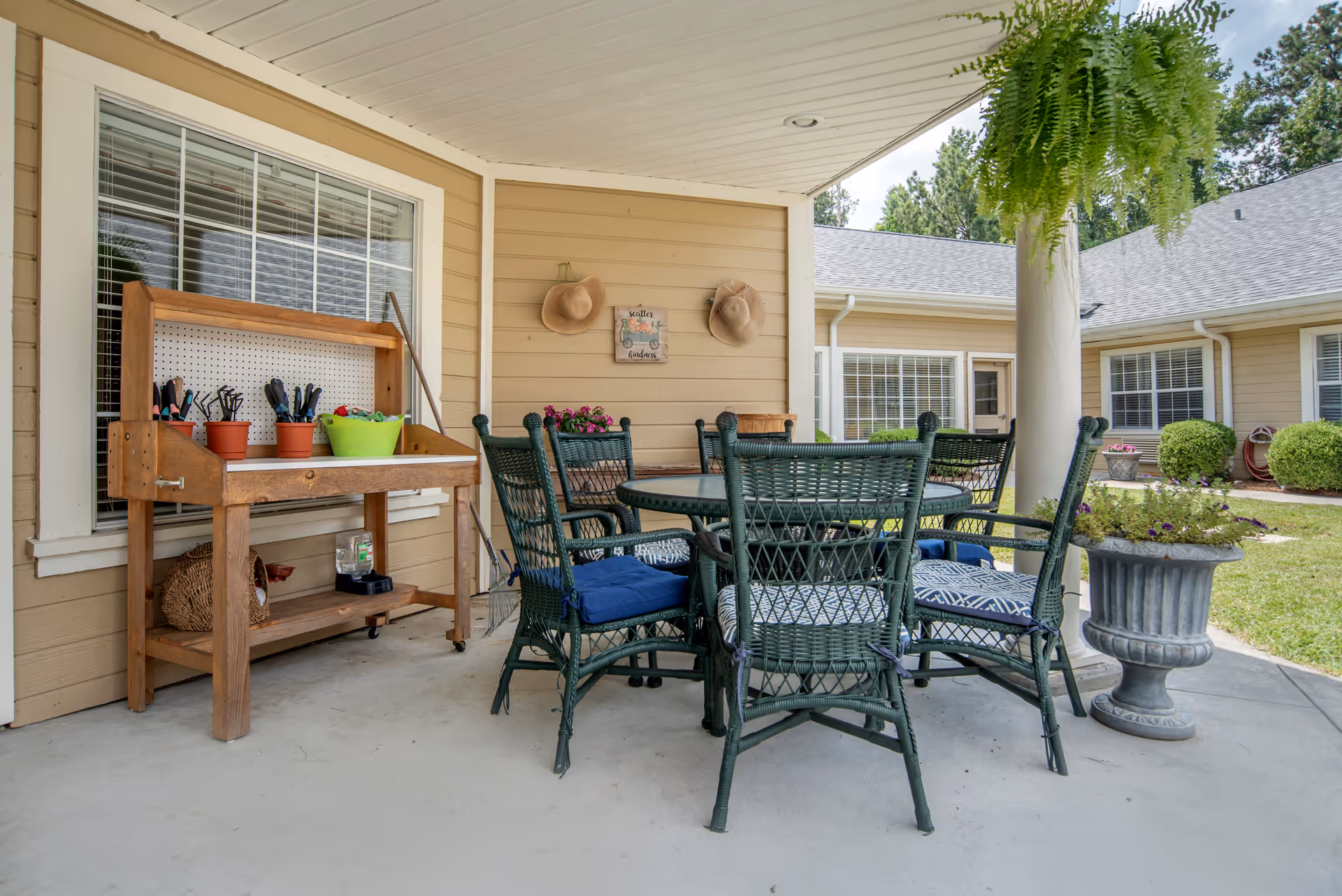Covered outdoor patio with a round wicker dining table and chairs, a potting bench with gardening tools, a hanging fern, and planters in front of a residential building.