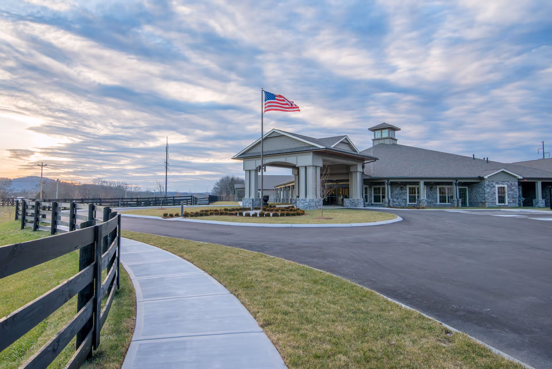 Driveway and covered entrance of a single-story senior living facility with an American flag, surrounding fence, and cloudy sky.