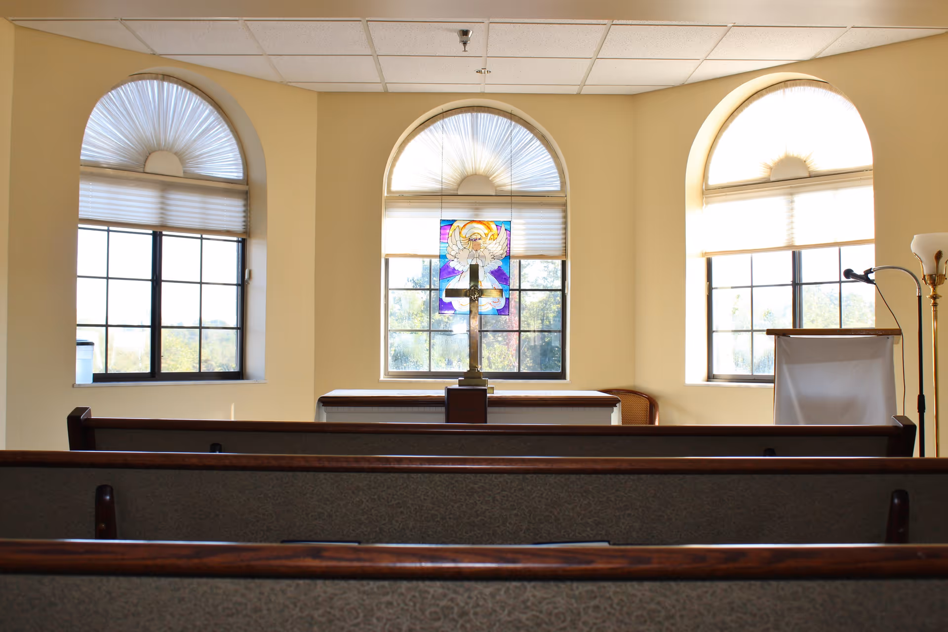 Interior view of a small chapel or prayer room with three arched windows, one featuring a stained glass angel. Wooden pews are arranged facing a small altar with a cross, and there is a lectern and a floor lamp on the right side.