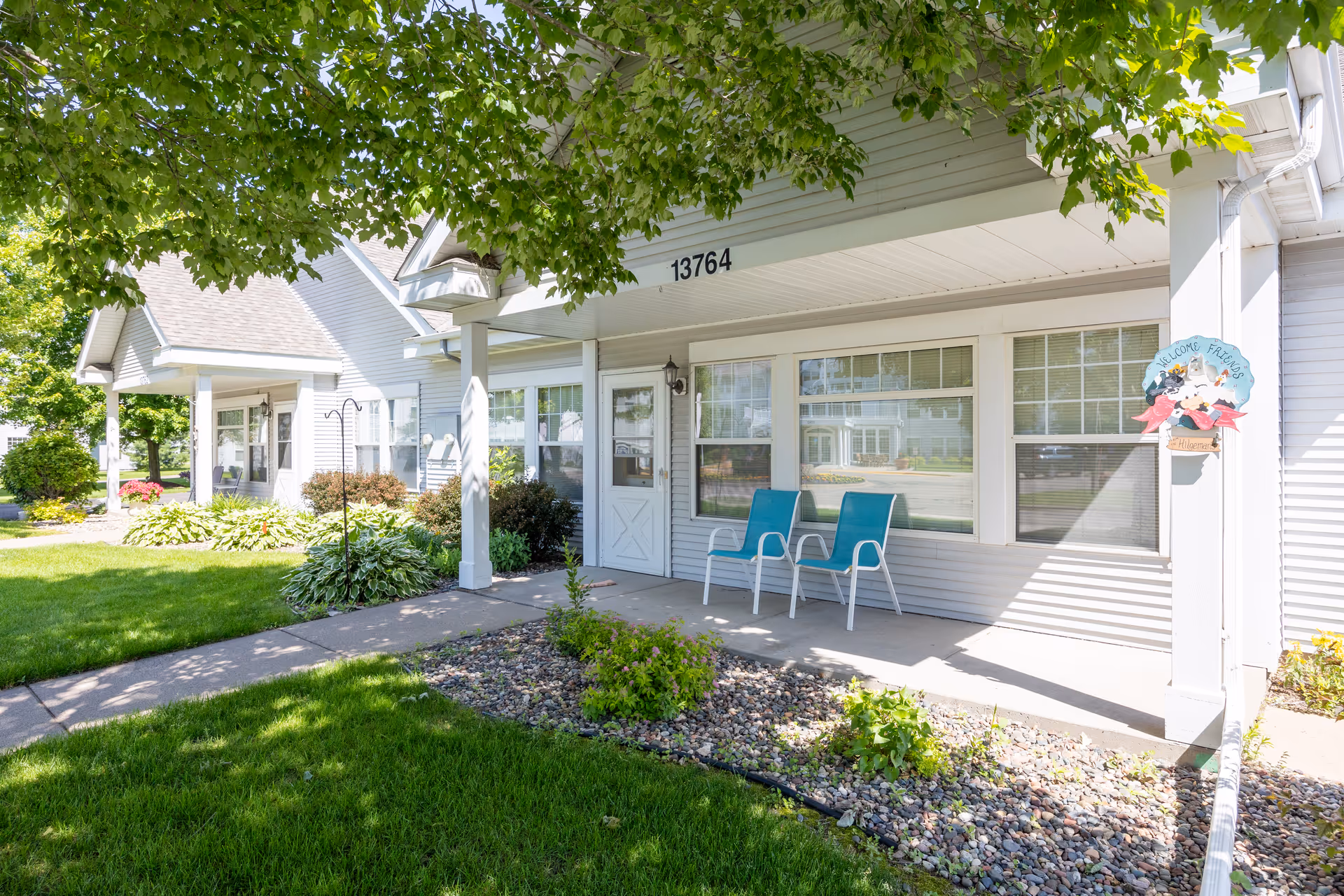 Front porch of a residential building at The Farmstead with two blue chairs, a white door, large windows, and a small garden bed with rocks and plants. The building number 13764 is visible above the porch, and a tree with green leaves provides shade.