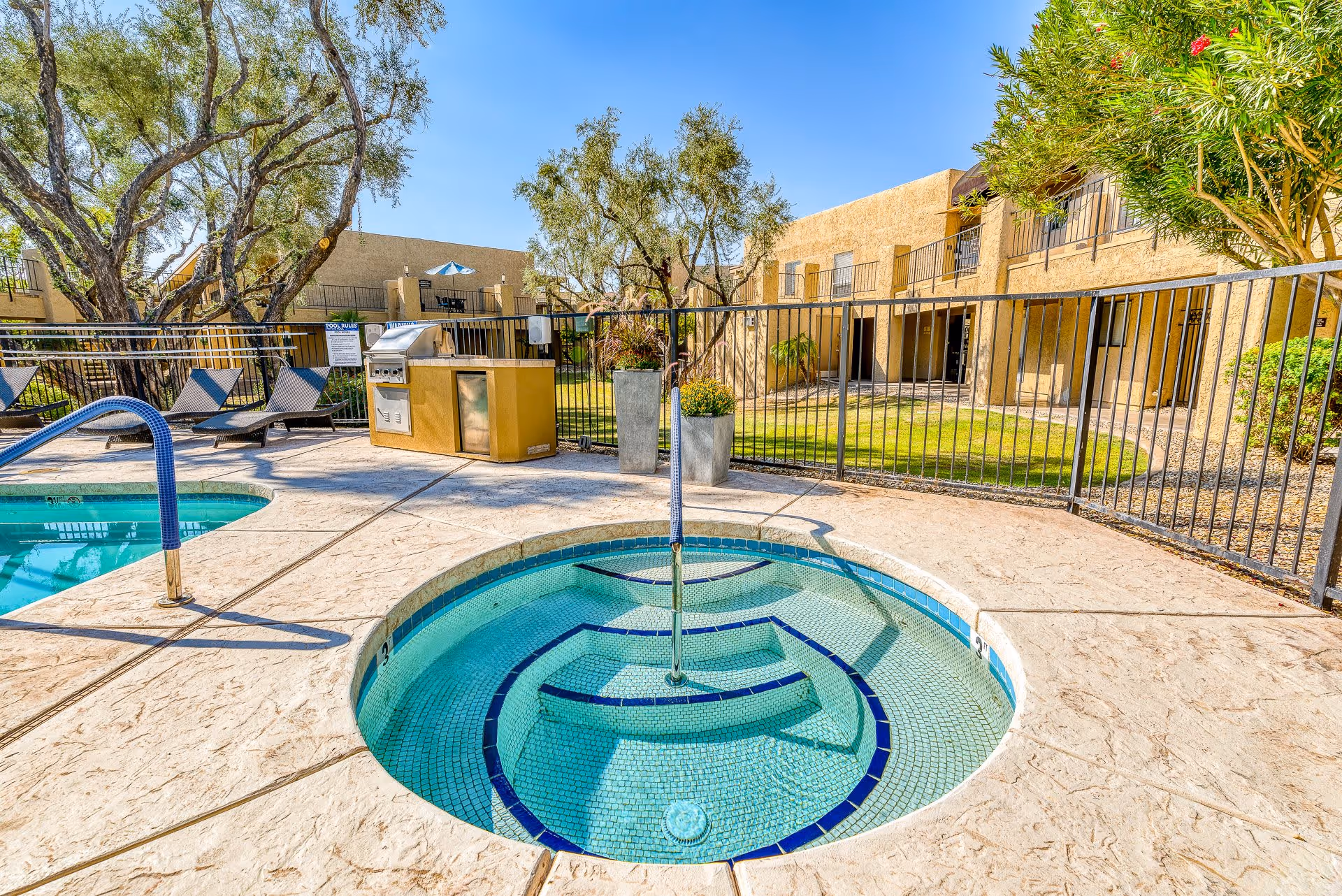 Outdoor area at Scottsdale Village Square featuring a small round hot tub with tiled steps and a metal handrail, adjacent to a swimming pool. The area is surrounded by a metal fence, with lounge chairs and a barbecue grill nearby. Trees and shrubs are visible, along with a two-story beige building in the background under a clear blue sky.