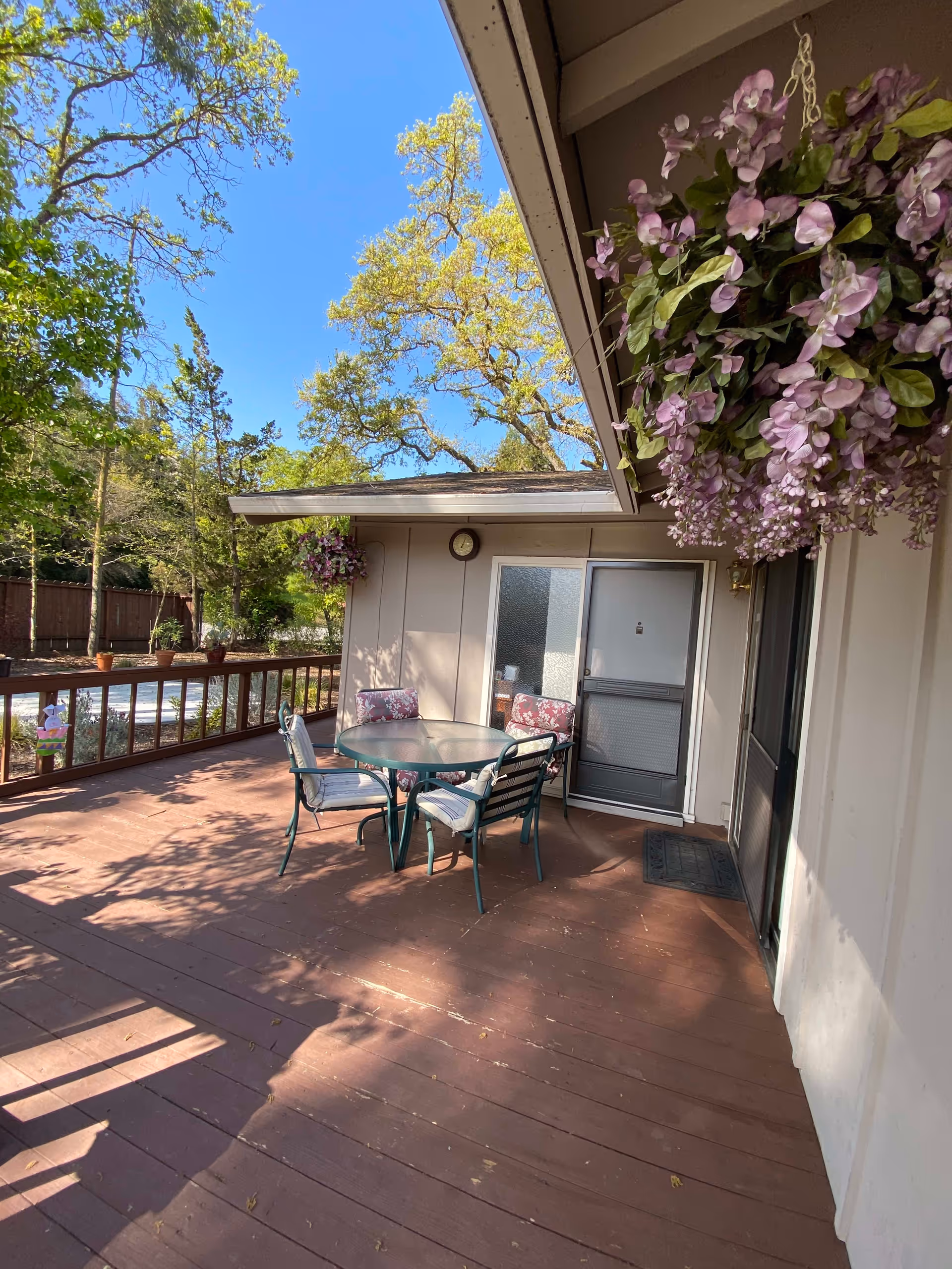 Outdoor wooden deck area with a round glass table and four chairs with cushions. There are hanging purple flowers on the right side and a clock mounted on the wall above a door. Trees and a wooden fence are visible in the background under a clear blue sky.