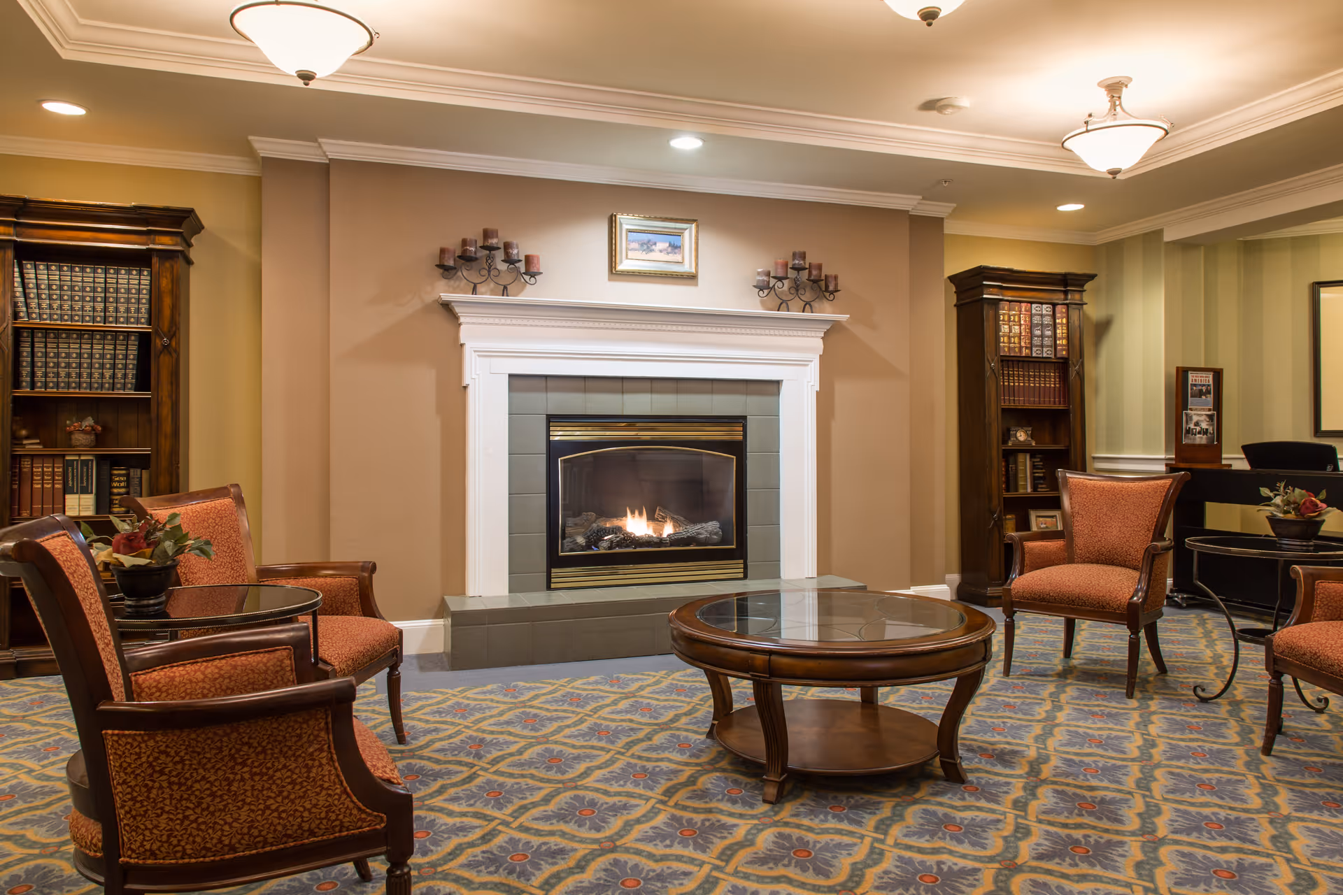 A cozy living room area with a lit fireplace centered on a beige wall. The room features four upholstered armchairs with wooden frames arranged around a round wooden coffee table with a glass top. Two tall wooden bookshelves filled with books flank the fireplace. The carpet has a patterned design in blue, yellow, and red tones. The ceiling has recessed lighting and two ceiling light fixtures.
