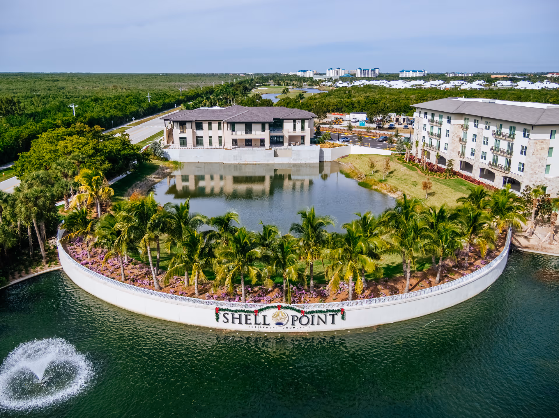 Aerial view of Shell Point Welcome Center showing a curved waterfront area with palm trees and landscaping, a water fountain in the pond, and buildings in the background surrounded by greenery and roads.