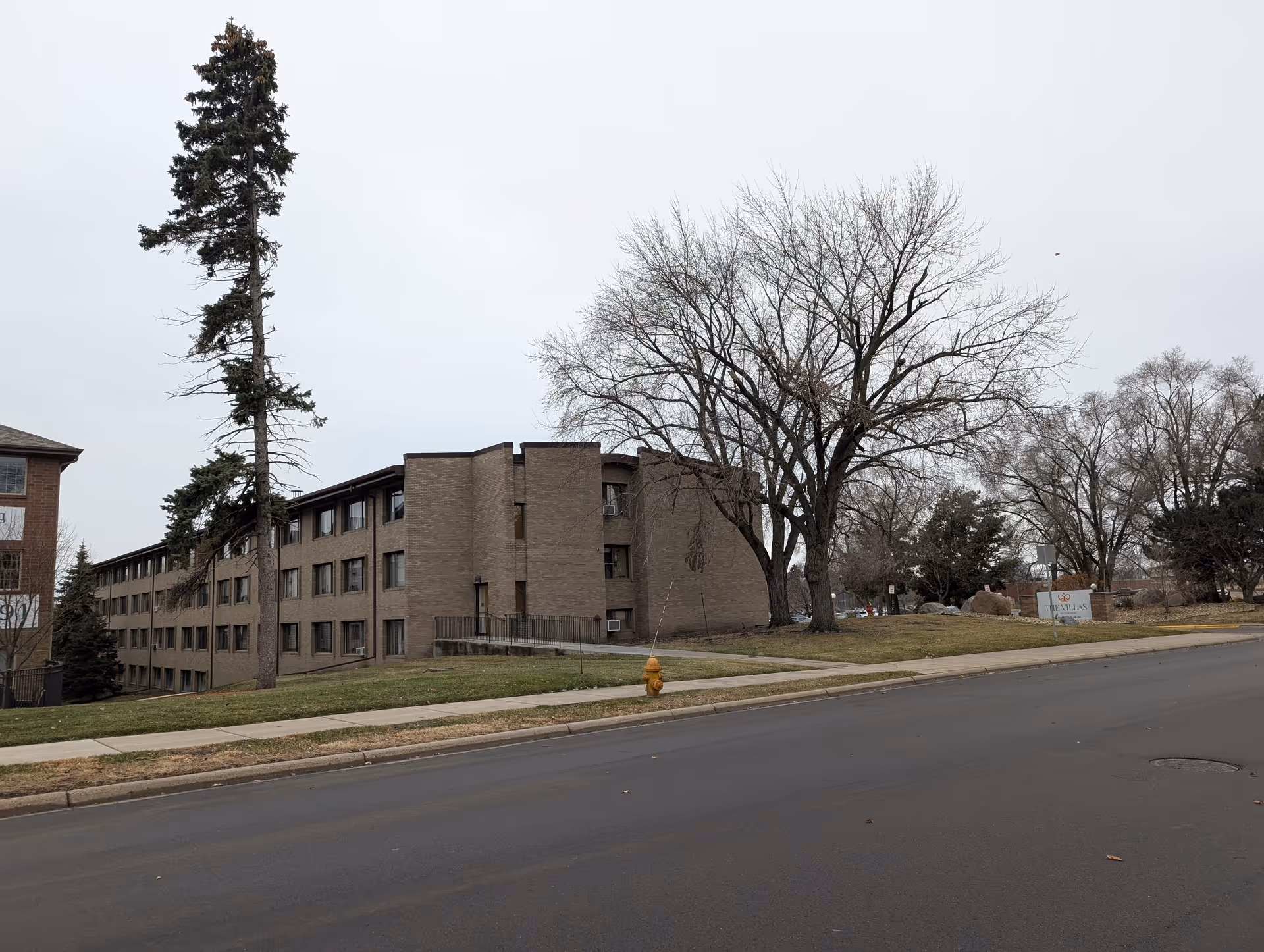 Exterior view of a multi-story senior living facility building with brown brick walls, several windows, a sidewalk, a fire hydrant, and leafless trees on a cloudy day.