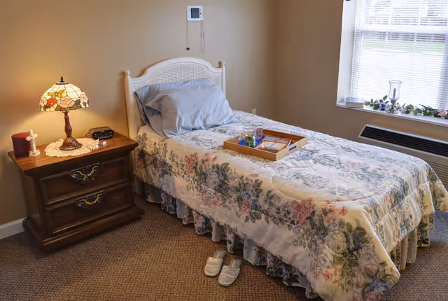 Single bed with a floral bedspread in a cozy bedroom beside a wooden nightstand, lamp, slippers, and a window with blinds.
