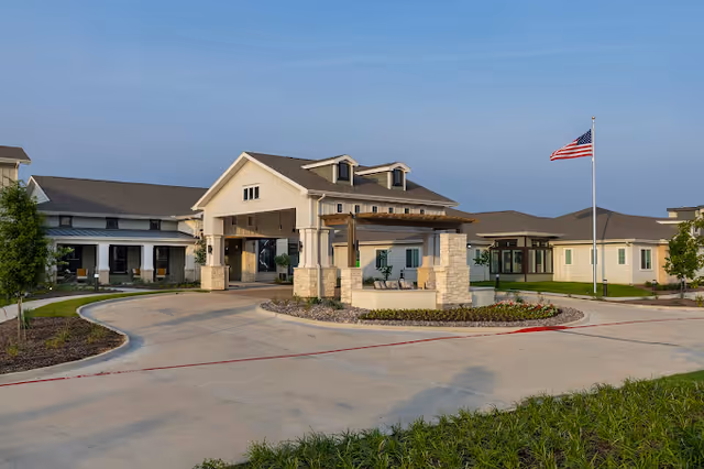 Exterior view of The Preserve at Spring Creek facility showing a large driveway with a covered entrance, landscaped areas, and an American flag on a flagpole under a clear sky.