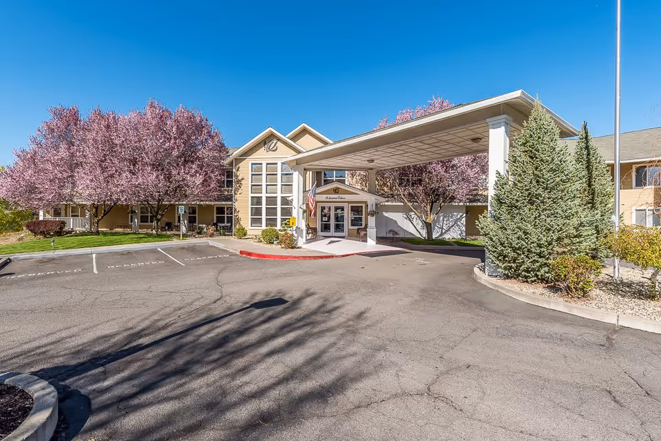 Exterior view of Klamath Falls Senior Living facility with a covered entrance, parking spaces, and blooming pink trees under a clear blue sky.