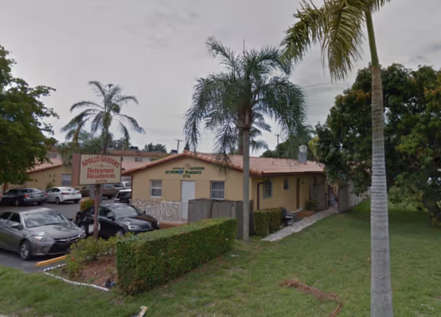 Exterior view of Apollo Gardens Retirement Residences, a single-story building with a beige exterior and a red-tiled roof, surrounded by palm trees and greenery. Several parked cars are visible in the parking lot next to the building. A sign near the entrance reads 'Apollo Gardens Retirement Residences'.
