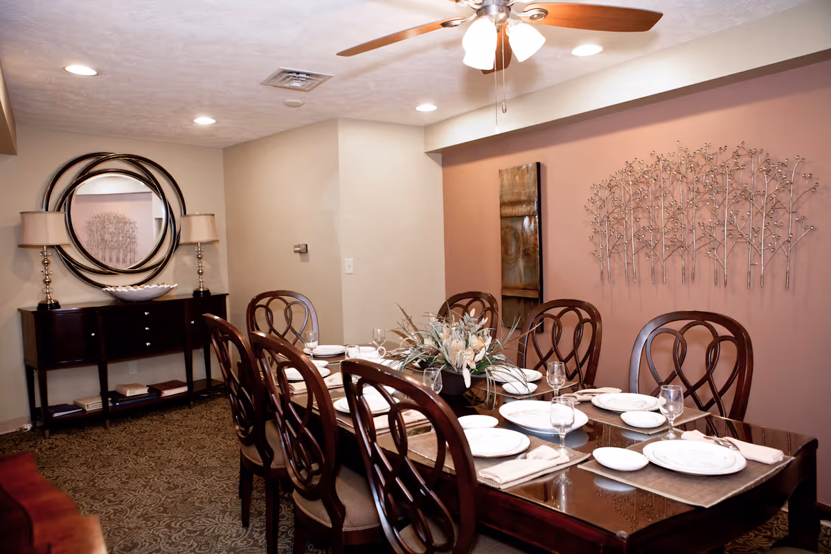 A formal dining room with a long wooden table set for eight people with white plates, napkins, and glassware. The room features a ceiling fan with lights, a decorative metal wall art piece, a tall rectangular mirror, and a sideboard with two lamps and a round mirror above it. The walls are painted in neutral tones with one accent wall in a muted pink color.