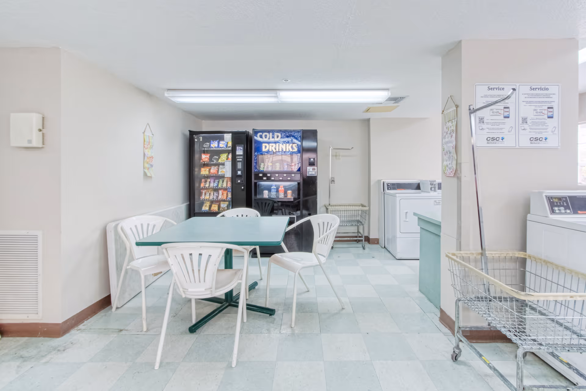 A laundry room with white washing machines and dryers along the right wall, a green table with four white plastic chairs in the center, and vending machines for snacks and cold drinks against the back wall. Laundry carts are positioned near the machines, and informational signs are posted on the walls.
