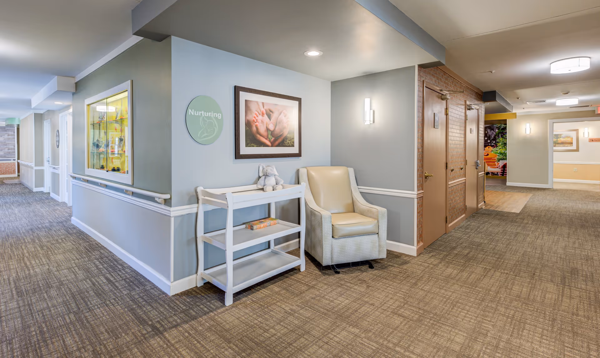 A well-lit hallway in a senior living facility with beige carpet and light blue walls. There is a white chair and a small white shelf with a stuffed elephant toy and a book. A framed picture of hands holding a small plant hangs on the wall above the chair. A round sign on the wall reads 'Nurturing'. The hallway has handrails and several doors, with a glimpse of a colorful outdoor area visible at the far end.