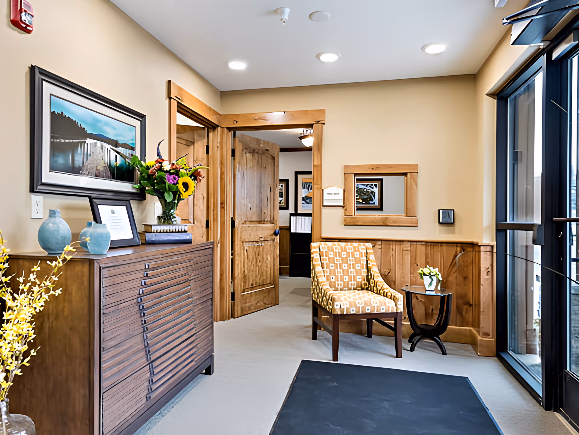 Welcoming interior entry area with wood trim, a patterned accent chair, decorative console with vases and flowers, and glass doors.