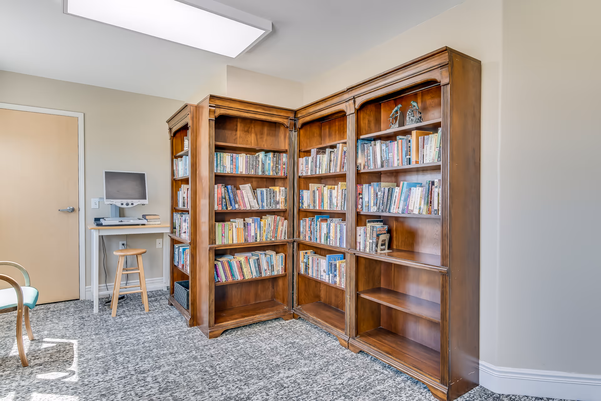 A cozy reading corner in a senior living facility featuring wooden bookshelves filled with books, a small desk with a computer monitor, a wooden stool, and a chair with light-colored upholstery. The room has a carpeted floor and neutral-colored walls.