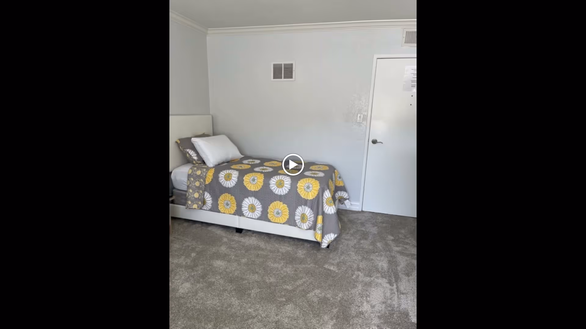 A simple bedroom with a single bed against a light gray wall. The bed has a white headboard and is covered with a gray bedspread featuring large white and yellow flower patterns. There are two pillows on the bed, one white and one gray. The floor is carpeted in a light gray color. A white door with a silver handle is visible on the right side of the room.